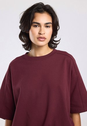 Jong persoon met middellang donker haar draagt een loszittend bordeauxrood T-shirt met korte mouwen, neutrale gezichtsuitdrukking, tegen een lichtgrijze achtergrond.