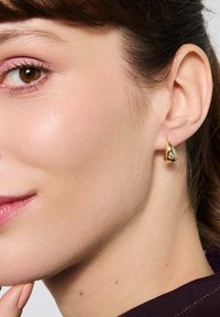 Złoty kolczyk w kształcie kropli z gładką powierzchnią i małym przezroczystym akcentem, noszony na małżowinie usznej, ukazujący bliski profil ucha.