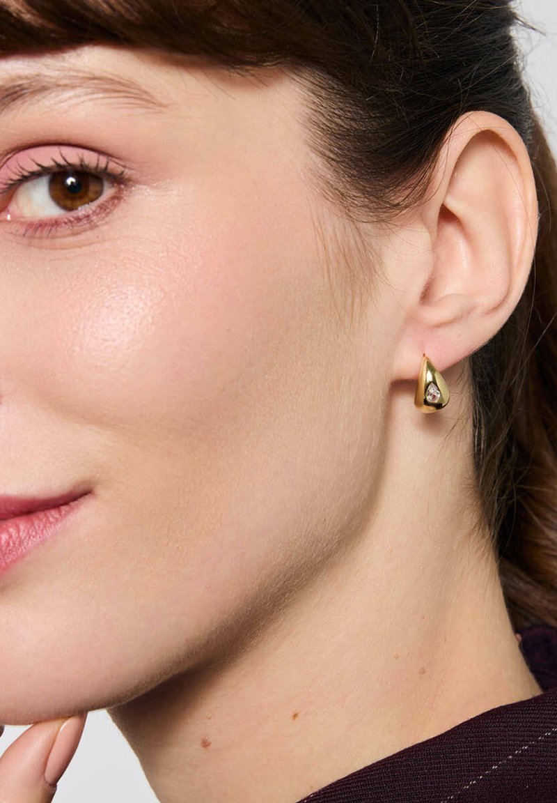 Złoty kolczyk w kształcie kropli z gładką powierzchnią i małym przezroczystym akcentem, noszony na małżowinie usznej, ukazujący bliski profil ucha.