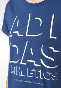 Blå bomull t-shirt med stora vita bokstäver som säger "ADIDAS ATHLETICS" med en slogan under. Korta ärmar och rundad halsringning.