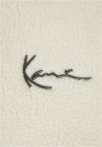 Kermanvärinen struktuurinen fleece-kangas, jossa on musta logo. Logo sisältää sulavia, kaarevia viivoja, joissa on pieni korostus.