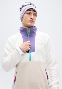 Πουλόβερ από φλις με λευκό σώμα, μωβ γιακά και μπεζ κάτω πάνελ. Διαθέτει τυρκουάζ φερμουάρ και μωβ/ροζ λεπτομέρειες στις τσέπες.