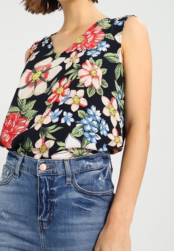Femme portant un haut noir sans manches avec un imprimé floral rouge, rose, blanc et bleu, rentré dans un jean bleu en denim taille haute.