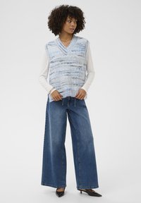 Göndör hajú nő kék és fehér kötött ujja nélküli pulóverben, ami egy fehér hosszú ujjú ing felett van, széles szárú farmernadrágban és fekete magassarkú cipőben.