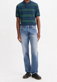 Man med en marinblå och grön randig polo, ljusblå raka jeans och svarta läderskor som står mot en vit bakgrund.