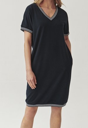 Kvinde iført en knælang sort kjole med hvide stribede kanter ved V-hals, ærmer og skørt, stående med hænderne i lommerne.