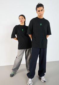 Svarte oversized t-skjorter med en liten grønn geometrisk logo, kombinert med løse grå og mørkeblå vide bukser. Teksturert bomullsstoff.