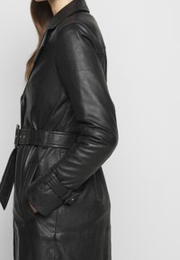 Black leather coat with a belted waist, side pockets, and a notched collar. Features long sleeves and a smooth, textured finish.