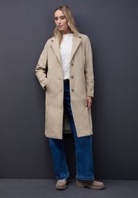 Beige Wollmischungsmantel mit einem Reverskragen, zwei vorderen Taschen und Knopfverschluss. Kombiniert mit einem weißen Strickpullover und blauen Schlagjeans.