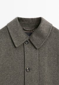 Cappotto grigio in lana con colletto classico, dotato di chiusura con bottone e una finitura morbida e testurizzata. Design semplice e colore neutro.