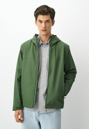 Junger Mann mit dunklen Haaren, der eine grüne Kapuzenjacke über einem grauen Hemd mit Knöpfen und hellblauen Jeans vor einem schlichten weißen Hintergrund trägt.
