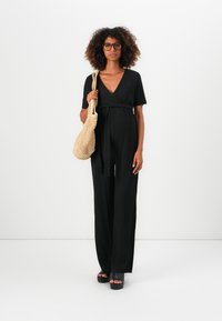 Kvinne med krøllete hår som har på seg svart jumpsuit, svarte plattformsandaler, briller og bærer en beige vevd skulderveske foran hvit bakgrunn.