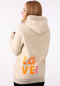 Haut à capuche beige avec cordon, arborant un audacieux texte "LOVE!" en orange et rose au dos. Tissu doux avec des poignets et un ourlet côtelés.