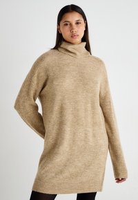Bezs színű, oversized garbó pulóver ribbelt ujjal és puha textúrával, amely laza szabással rendelkezik, és a comb közepéig ér.