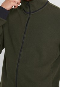 Grüne Reißverschlussjacke aus strukturiertem Stoff mit hohem Kragen und marineblauen Akzenten an den Bündchen und dem Reißverschluss.