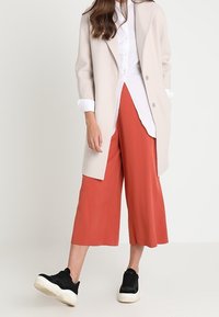 Béžový dlouhý kabát, bílá košile na knoflíky a kalhotová sukně barvy rezavé. Černé tenisky s bílými podrážkami. Hladké textury a uvolněný střih.