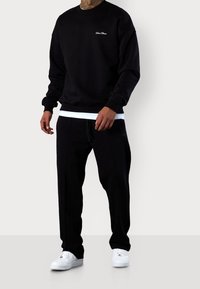Uomo con felpa nera e pantaloni jogger abbinati, indossando sneakers bianche, in piedi su uno sfondo grigio.