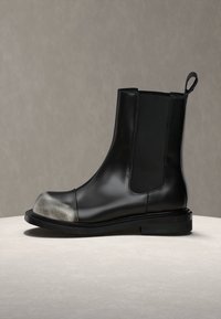 Bottine noire en cuir avec des panneaux latéraux élastiques et un bout métallique argenté, présentée sur une surface claire sur un fond neutre.