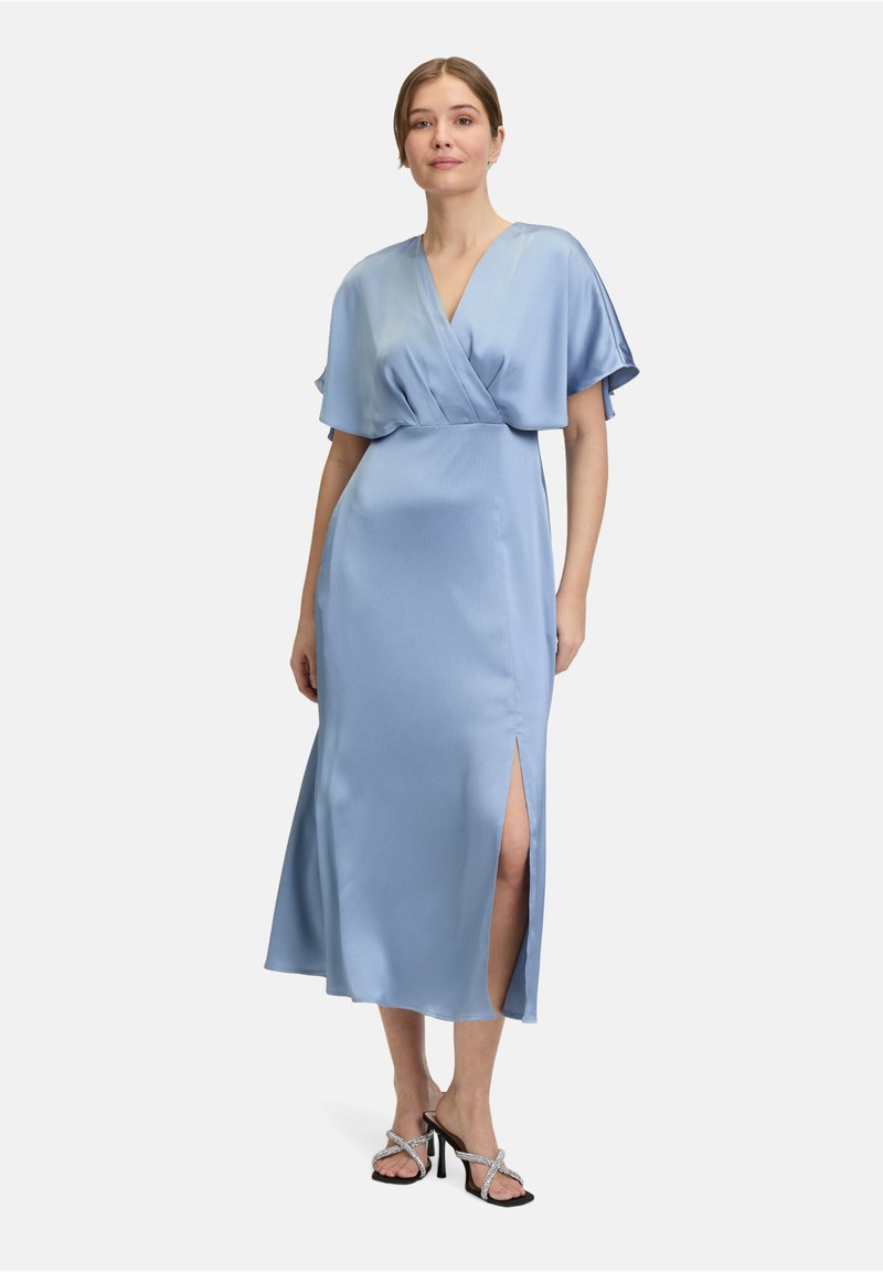 Robe en satin bleu clair avec un design croisé, manches courtes et volant, et une fente sur le côté gauche. Associée à des sandales à talons noires.