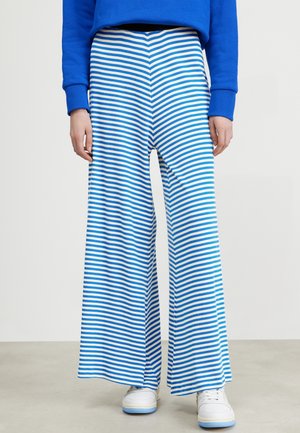 Osoba ubrana w niebiesko-białe spodnie w szerokie paski, niebieską bluzę i białe trampki stojąca na szarej podłodze.