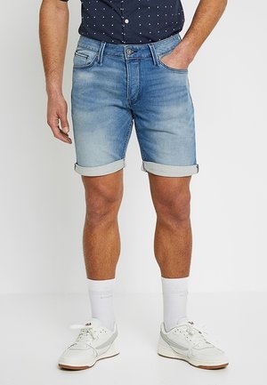 Pantalones cortos de mezclilla azul claro con un ajuste regular, dobladillos enrollados, cinco bolsillos y cierre de botón en el frente. Combinados con zapatillas blancas y calcetines.