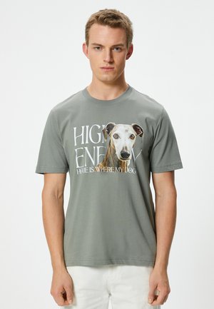 Camiseta gris de manga corta con un gran gráfico de perro y el texto "EN CASA CON ALTA ENERGÍA ES DONDE ESTÁ MI PERRO". Material de algodón suave, ajuste relajado.