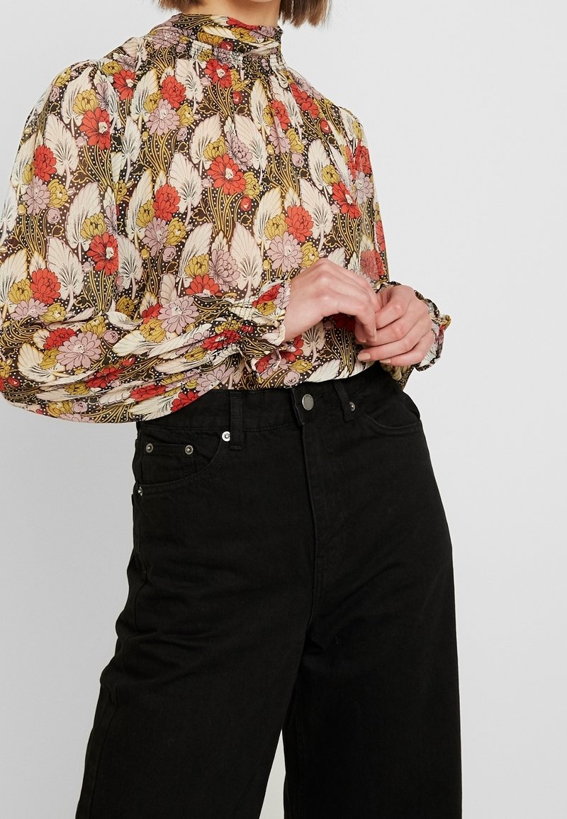 Langärmlige Bluse mit floralen Mustern in Creme-, Rot- und Gelbtönen; geraffter Ausschnitt und Bündchen; kombiniert mit hochgeschnittenen schwarzen Jeans.