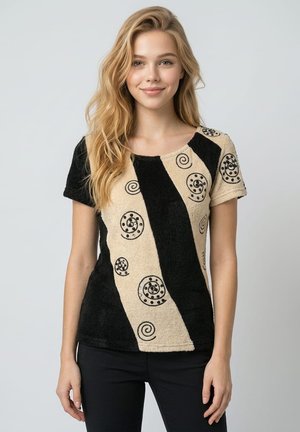 Joven mujer con cabello rubio largo, vestida con una camisa de manga corta negra y beige con patrones en espiral y pantalones negros, de pie frente a un fondo gris.