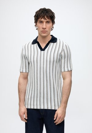 Homme aux cheveux courts et bouclés portant un pull en maille blanc à manches courtes avec des rayures verticales noires et un col noir, debout devant un fond uni.