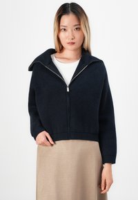 Ung kvinne med rett blondt hår iført en marineblå hettegenser over en hvit topp og beige skjørt mot en hvit bakgrunn.