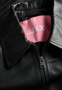 Fekete bőrdzseki elülső cipzárral, nagy gallérral és belül jól látható rózsaszín címkével, amelyen a "JJXX" márkanév olvasható.