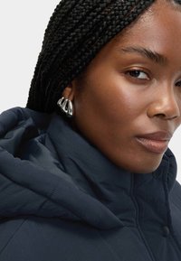 Mørkeblå pufferjakke med høj krave, der har en glat tekstur. Sølvfarvede hoops øreringe fremhæver personens ansigt.