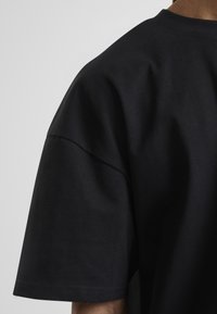 Acercamiento de una persona que lleva una camiseta negra de manga corta lisa, mostrando el hombro y la parte superior del brazo contra un fondo gris claro.