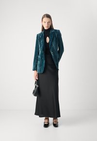 Teal velvet blazer with vertical stripes, layered over a black long-sleeve dress, holding a small black handbag, matched with black shoes.