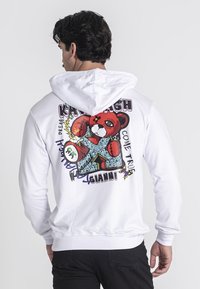 Sudadera blanca con un gráfico de oso de dibujos animados colorido en la parte posterior, con elementos de texto y contornos gruesos. Material de mezcla de algodón, corte relajado.