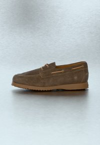 Mocassin marron en daim à enfiler avec des lacets beiges et une semelle en caoutchouc sur un fond gris neutre.
