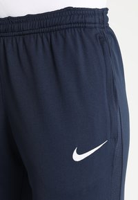 Marinblå atletiska byxor med elastisk midja, sidofickor och en vit Nike-logga på vänster lår. Slät tygtextur.