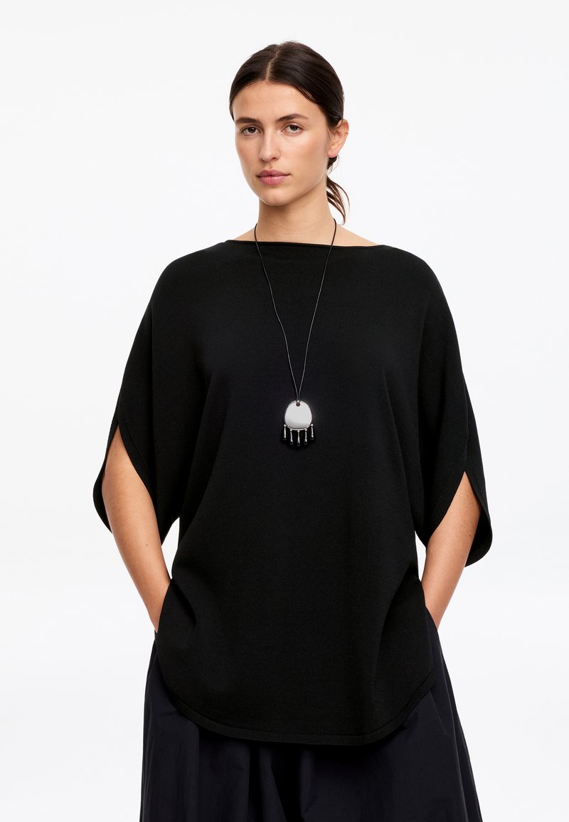 Femme portant un haut noir ample avec des manches aux coudes, mains dans les poches, et un long collier avec un pendentif rond et des perles pendantes.