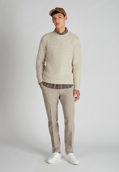Beige ribbeltrui, geruit shirt eronder, lichtbeige broek en witte sneakers. Model draagt een bruine pet. Eenvoudig, textuurontwerp.
