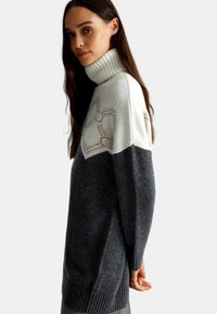 Maglione a collo alto in bianco e grigio scuro, con un design geometrico in colori chiari, dotato di polsini e bordo a coste per aggiungere texture.