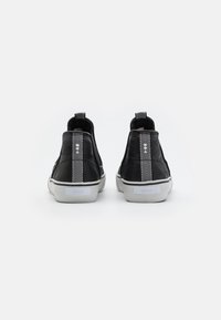 Baskets noires à enfiler avec semelles blanches, languettes arrière à motif chevron, placées côte à côte sur fond blanc uni.