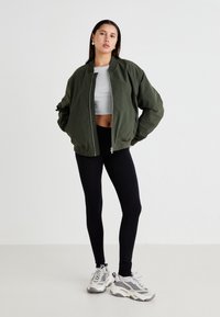 Olívazöld bombázó dzseki, cipzárral záródik, szürke crop top, fekete leggings és szürke, hálós részletekkel rendelkező sportcipő társaságában.