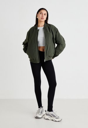 Olívazöld bombázó dzseki, cipzárral záródik, szürke crop top, fekete leggings és szürke, hálós részletekkel rendelkező sportcipő társaságában.