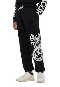 Czarne dresy z elastycznym pasem ze sznurkiem, z białym tekstem w stylu graffiti oraz nadrukiem uśmiechniętej buźki na lewej nodze. W połączeniu z białymi trampkami.