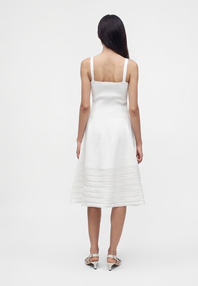 Femme aux longs cheveux foncés portant une robe midi blanche sans manches et des sandales blanches à talons, debout de dos sur un fond uni.