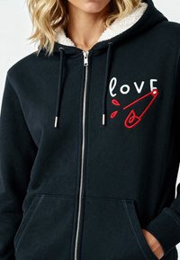 Hoodie bleu marine avec un doublure en fausse fourrure, zip, poches avant et design brodé "love" avec un accent de pince de sécurité rouge.