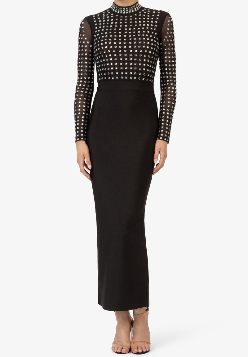 Robe longue à manches longues noire avec ornements carrés argentés sur le corsage et les manches, taille cintrée, jupe longueur cheville, portée avec des escarpins couleur nude.