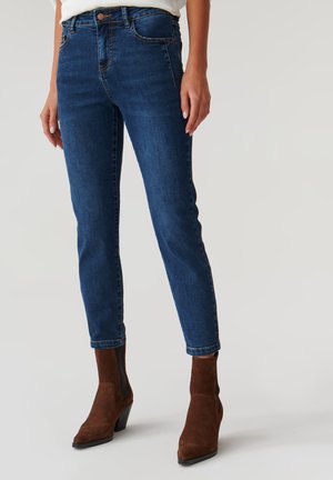 Dunkelblaue Denim-Jeans mit slim fit, ausgestattet mit klassischen Taschen und dezentem Nähen, kombiniert mit braunen Wildleder-Stiefeletten.