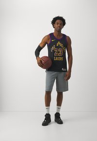 Čierna basketbalová dres Los Angeles Lakers s žltými akcentmi a číslom 23. Sivé športové šortky. Hráč drží basketbal a má na sebe čierne topánky.