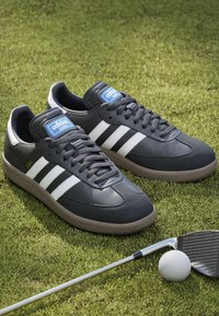 Czarne skórzane sneakersy z trzema białymi paskami, teksturowana gumowa podeszwa, niebieska metka na języku i zaokrąglony czubek, ustawione na zielonej trawie obok piłki golfowej i kija golfowego.